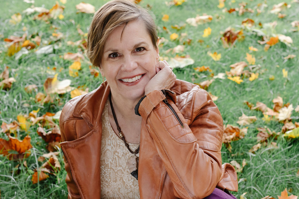 Karen Ann Bulluck resting on the grass in a tan leather jacket with fall leaves around.